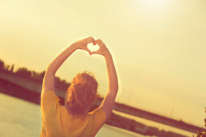 Woman making a heart with her arms stretched over her head making a heart with her hands looking over the water.